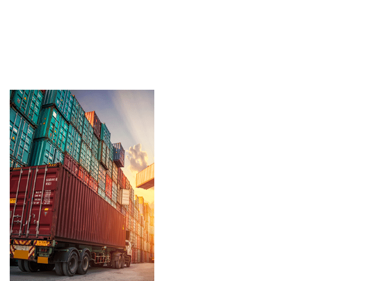 A semi-trailer truck carrying a shipping container drives past stacked containers at sunset.