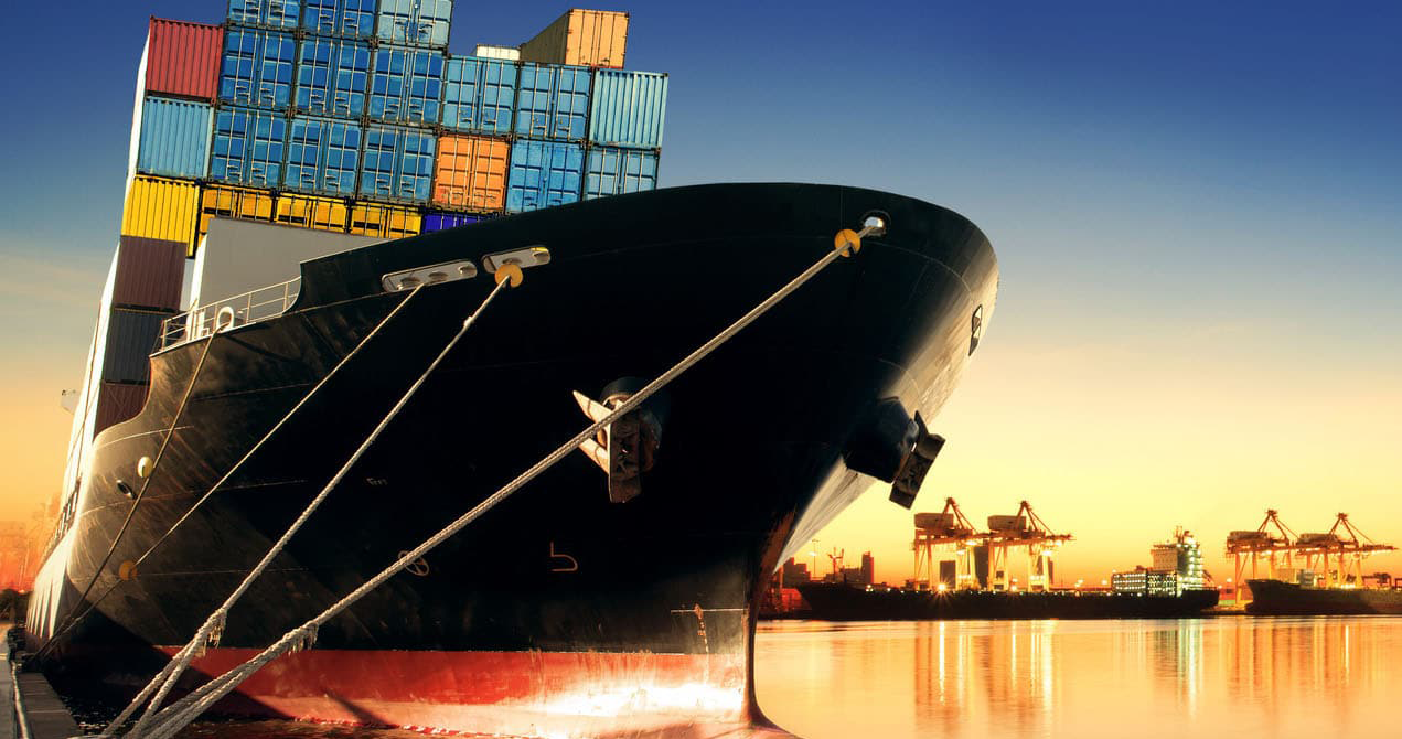 A loaded container ship docked at port with cranes in the background at sunset.