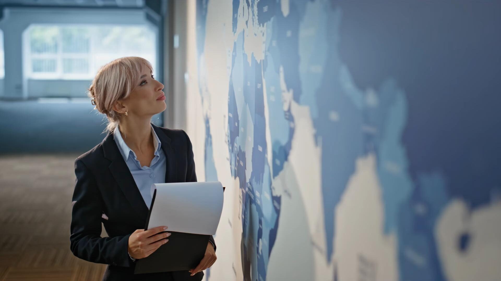 A businesswoman holding documents examines a large world map mounted on an office wall.