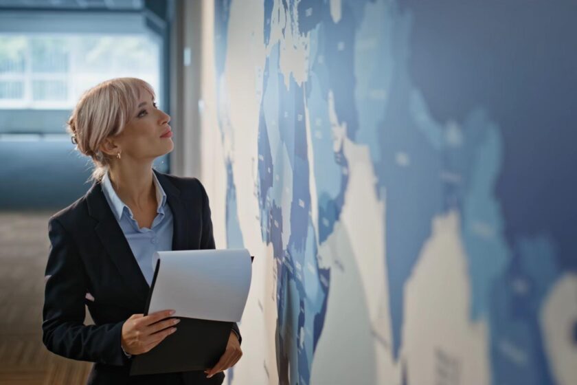 A businesswoman holding documents examines a large world map mounted on an office wall.